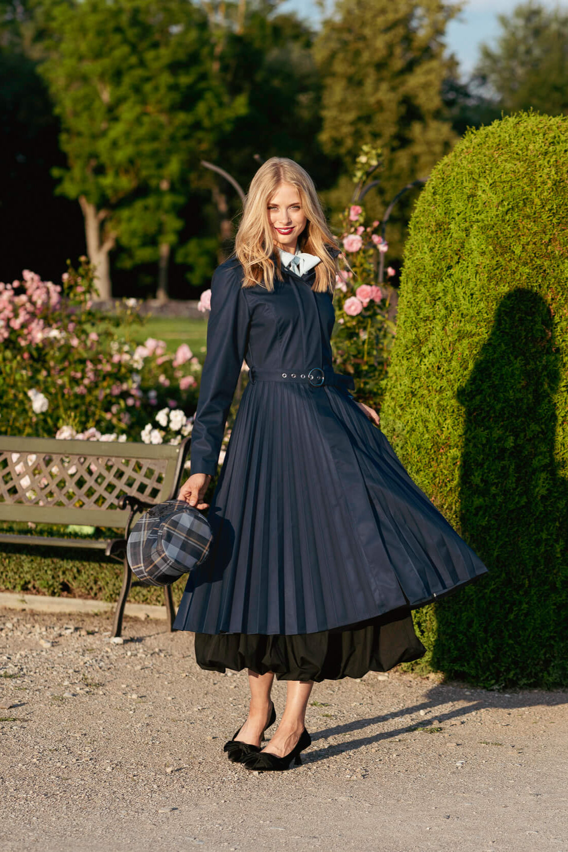 Woman in a long dark blue coat standing in a garden with flowers and trees.