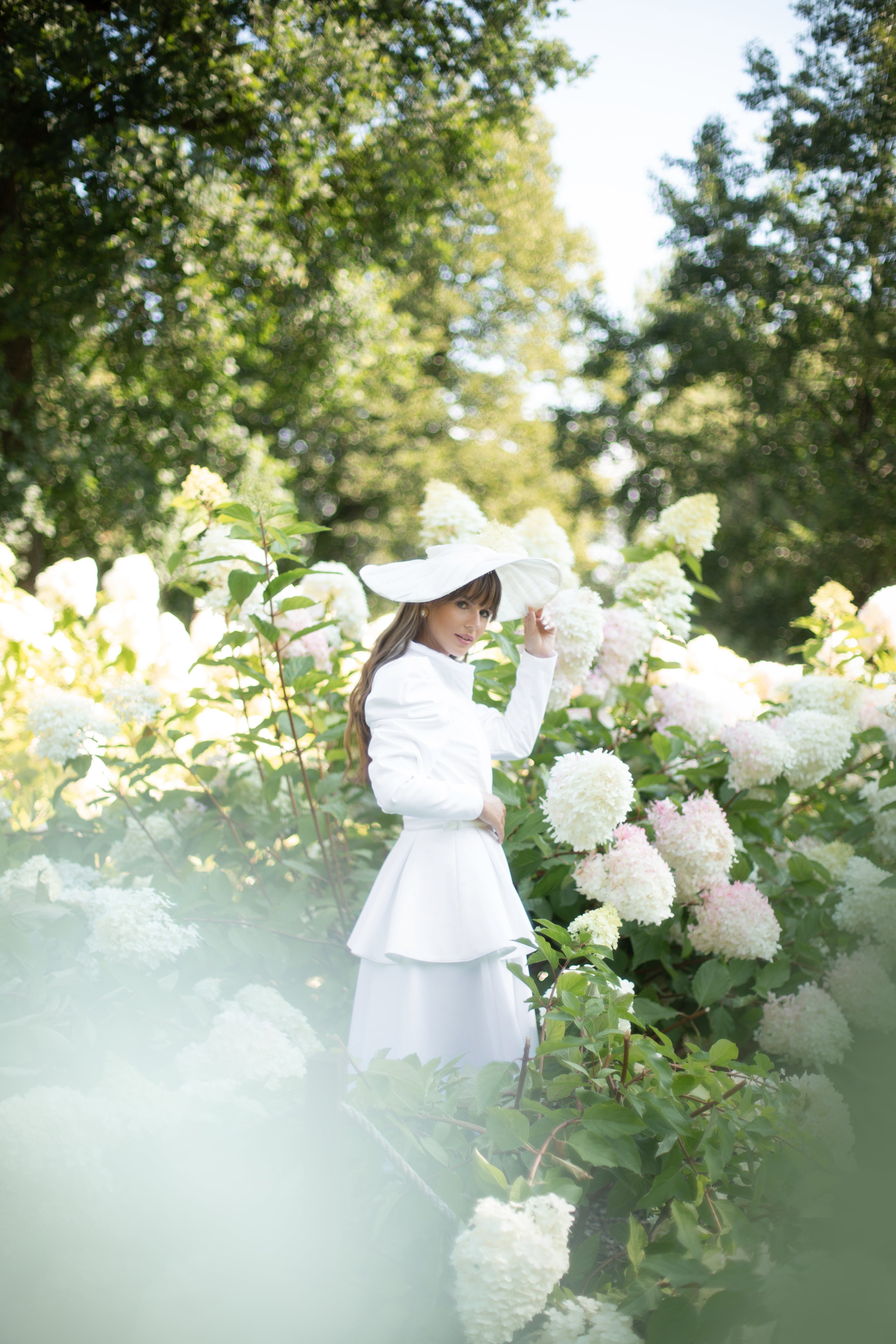 A woman in a white coat and matching wide-brim hat stands in a sunlit garden among white and pale pink hydrangea blooms, with green trees in the background.