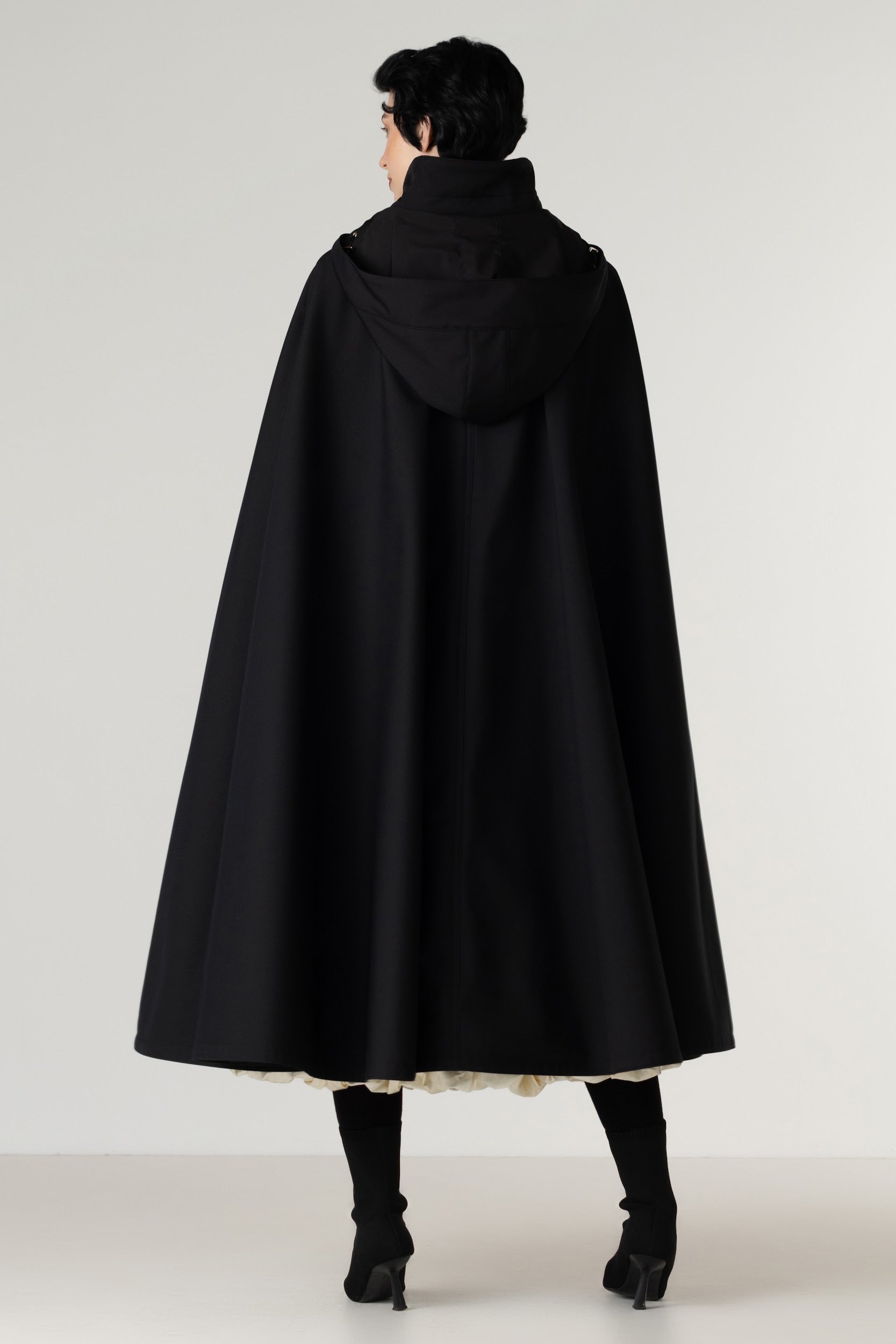 A model shown from the back wearing a black hooded cape coat by RainSisters, black tights, and black heels against a plain light gray studio background.