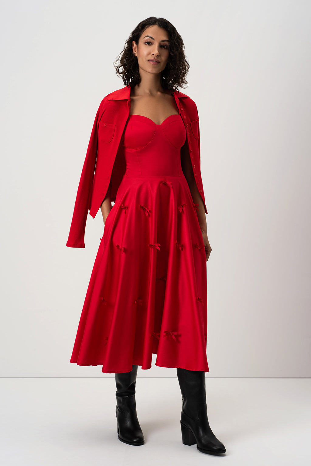 A woman modeling RainSisters bright red midi dress Little Red Bows with a sweetheart neckline, a matching structured jacket draped over her shoulders, and black ankle boots, photographed against a plain light gray studio background.