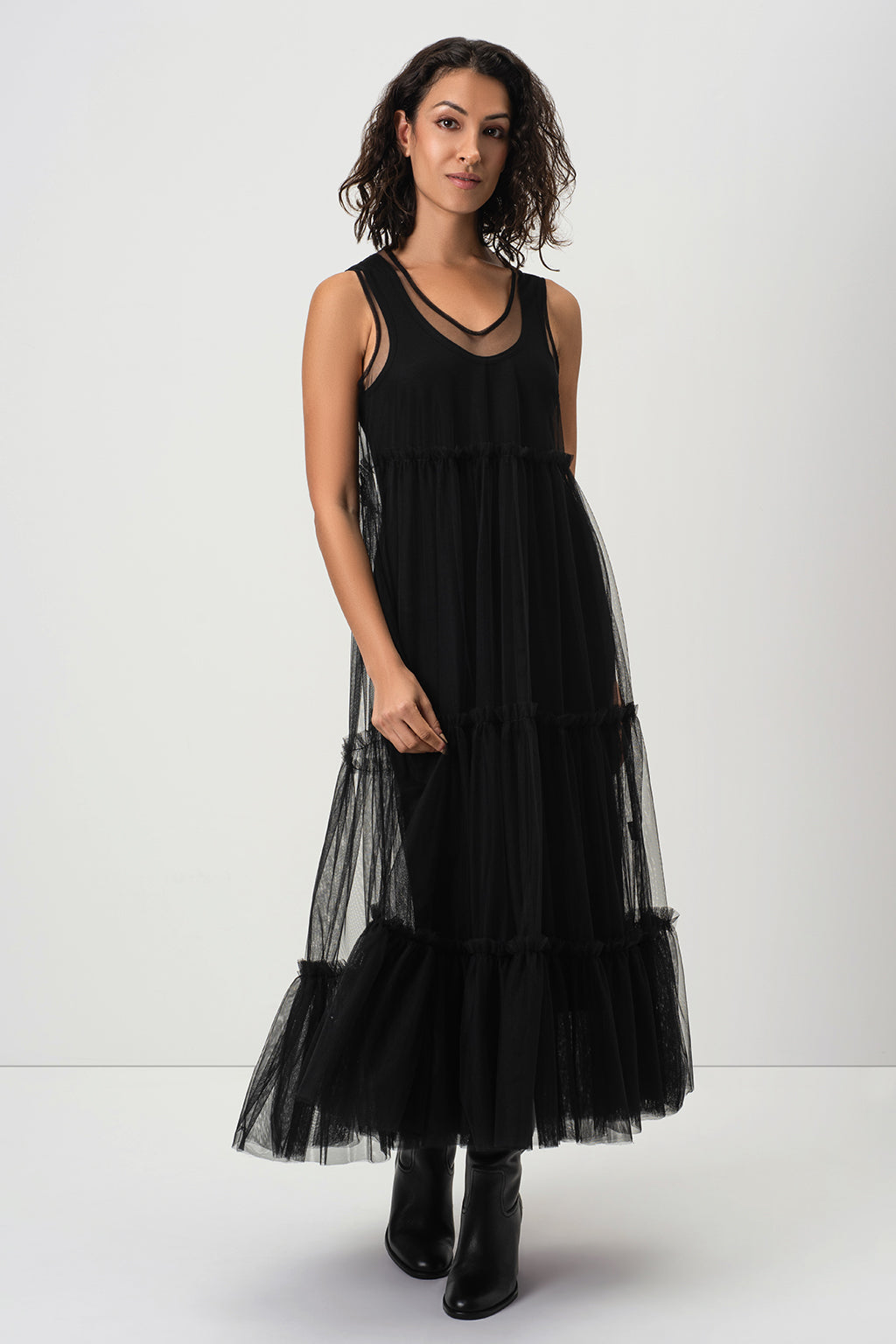 A woman modeling RainSisters sleeveless black tiered tulle maxi dress Sheer Romance with black ankle boots, standing against a plain light gray studio background.