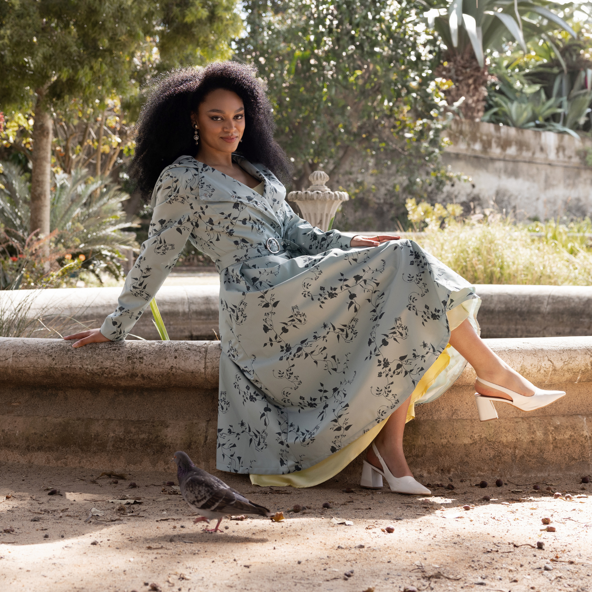 A woman with voluminous dark curly hair sits on a fountain ledge outdoors, wearing a light green coat Minty Meadow by RainSisters with a dark green floral print and white block-heel sandals.