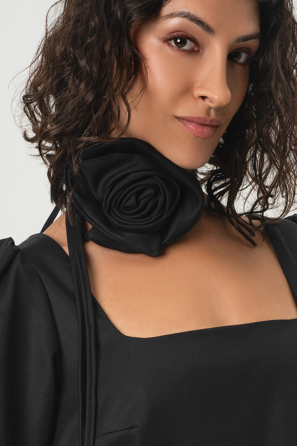 A close-up of a woman wearing RainSisters black satin rose collar choker with tie straps over a black dress, against a light gray background.