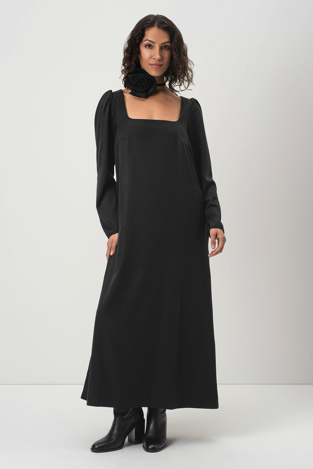 A woman modeling RainSisters long black dress Dark Muse with long sleeves, a square neckline, and a large black fabric flower detail, paired with black ankle boots against a light gray studio background.