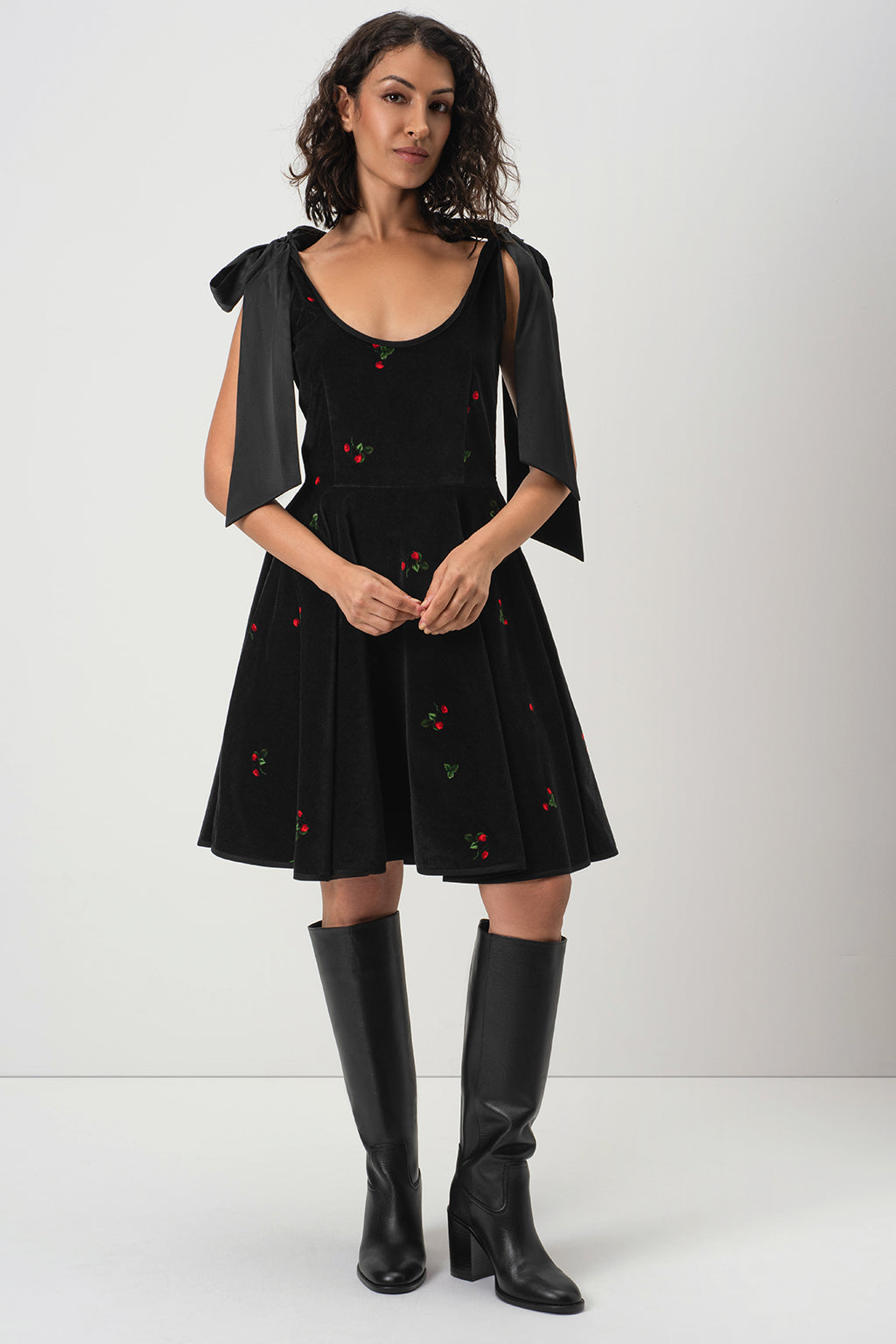 A woman models RainSisters black sleeveless velvet dress Berry Swing with satin bow shoulder details, a flared skirt, and small red strawberry motifs, paired with black knee-high boots against a light gray studio background.