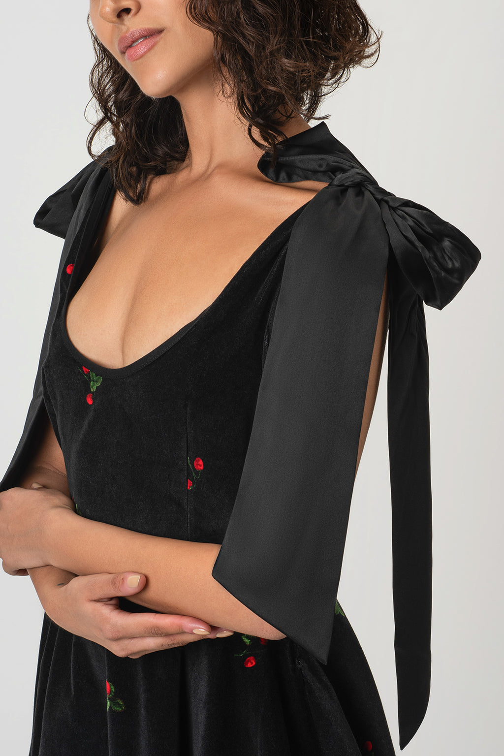 A close-up of a woman wearing RainSisters black velvet dress Berry Swing with a deep scoop neckline, wide draped shoulder ties, and small red embroidery.