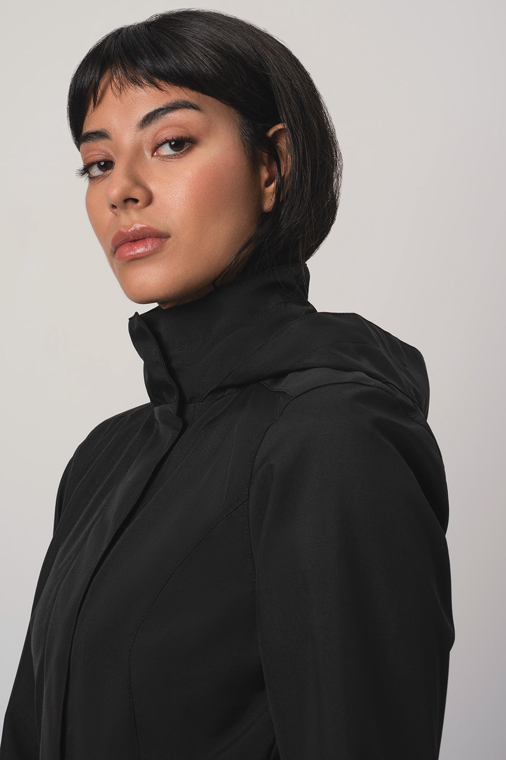 A woman with a dark hair wearing RainSisters black hooded jacket Black Sway, shown in a side profile against a light gray studio background.