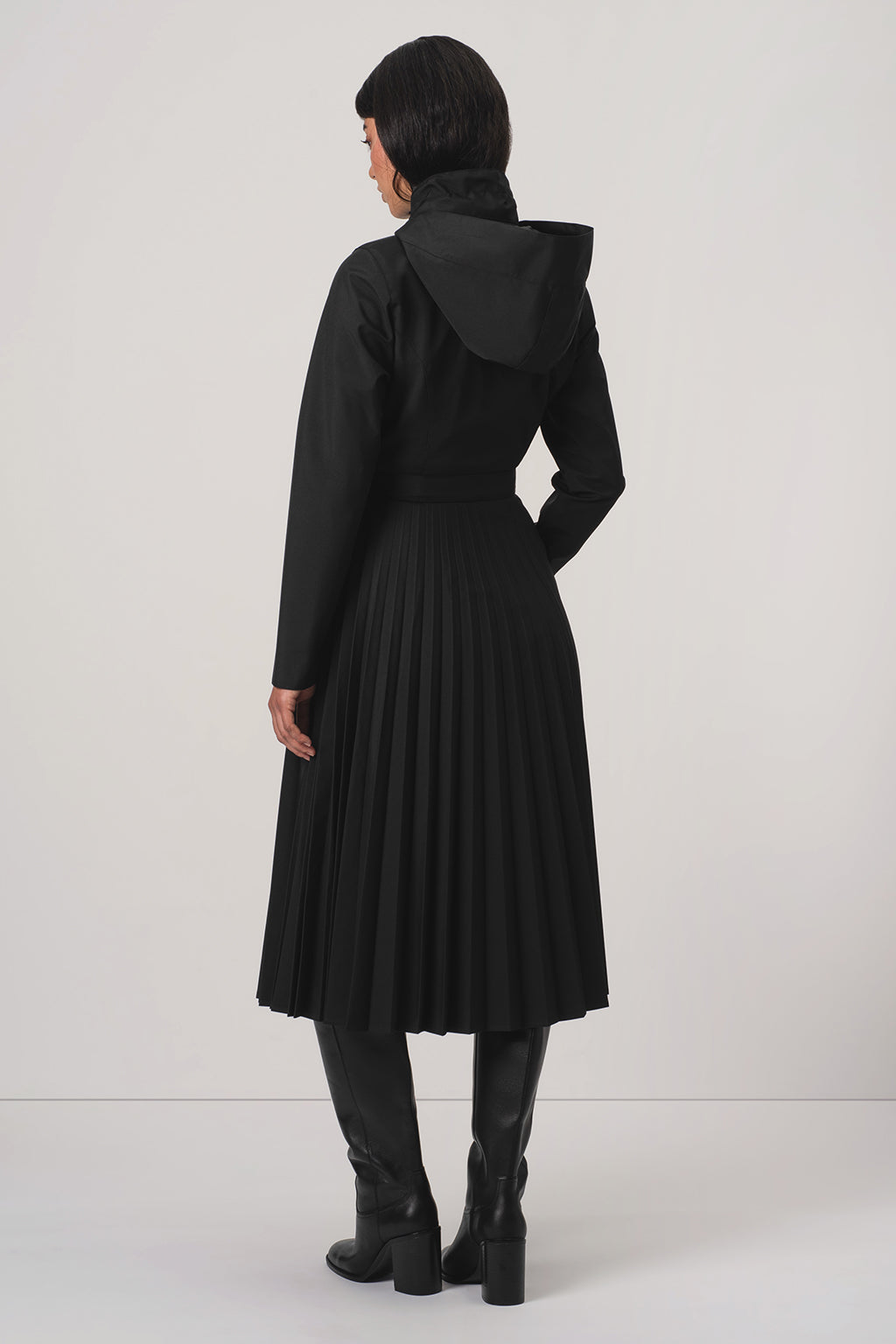 A model viewed from the back wearing RainSisters black hooded long-sleeve coat Black Sway with a pleated skirt and black knee-high boots, set against a plain light gray studio background.