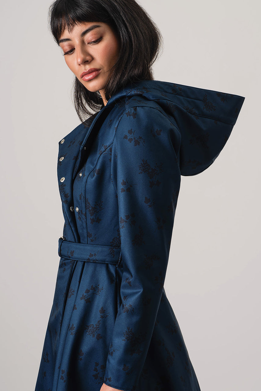 A woman wearing RainSisters dark navy hooded raincoat Blue Frost with a subtle dark floral pattern, photographed against a neutral light gray studio background.