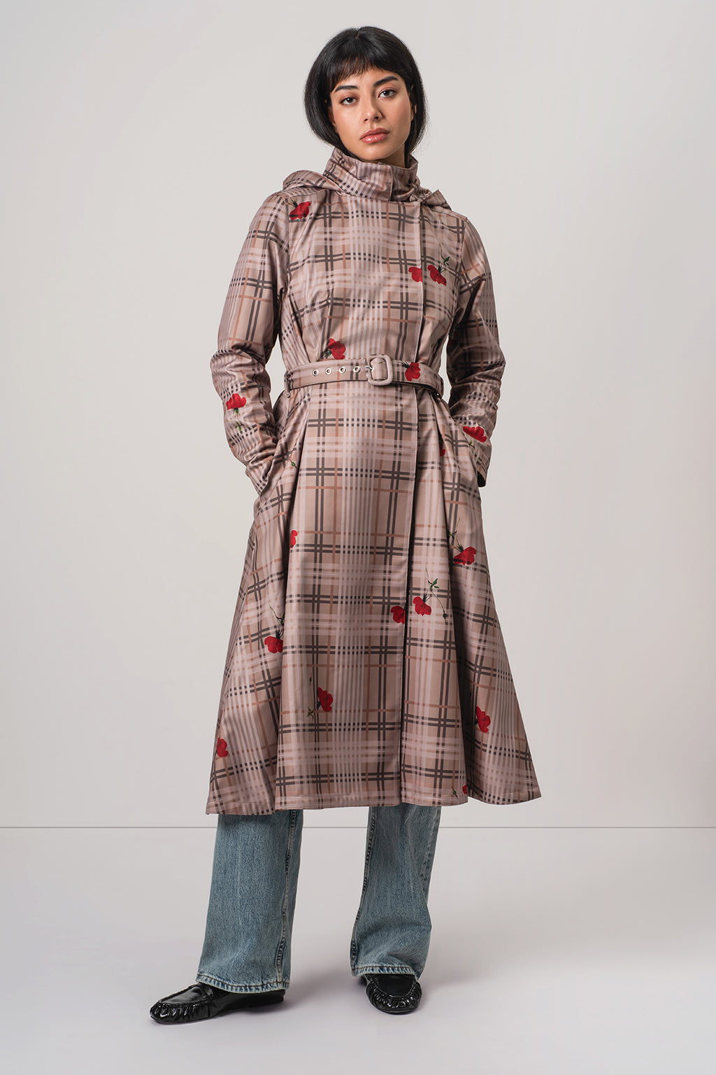 A model wearing RainSisters brown and beige plaid hooded coat Blush of the Crown with red flower motifs, styled with blue jeans and black shoes against a light gray studio background.