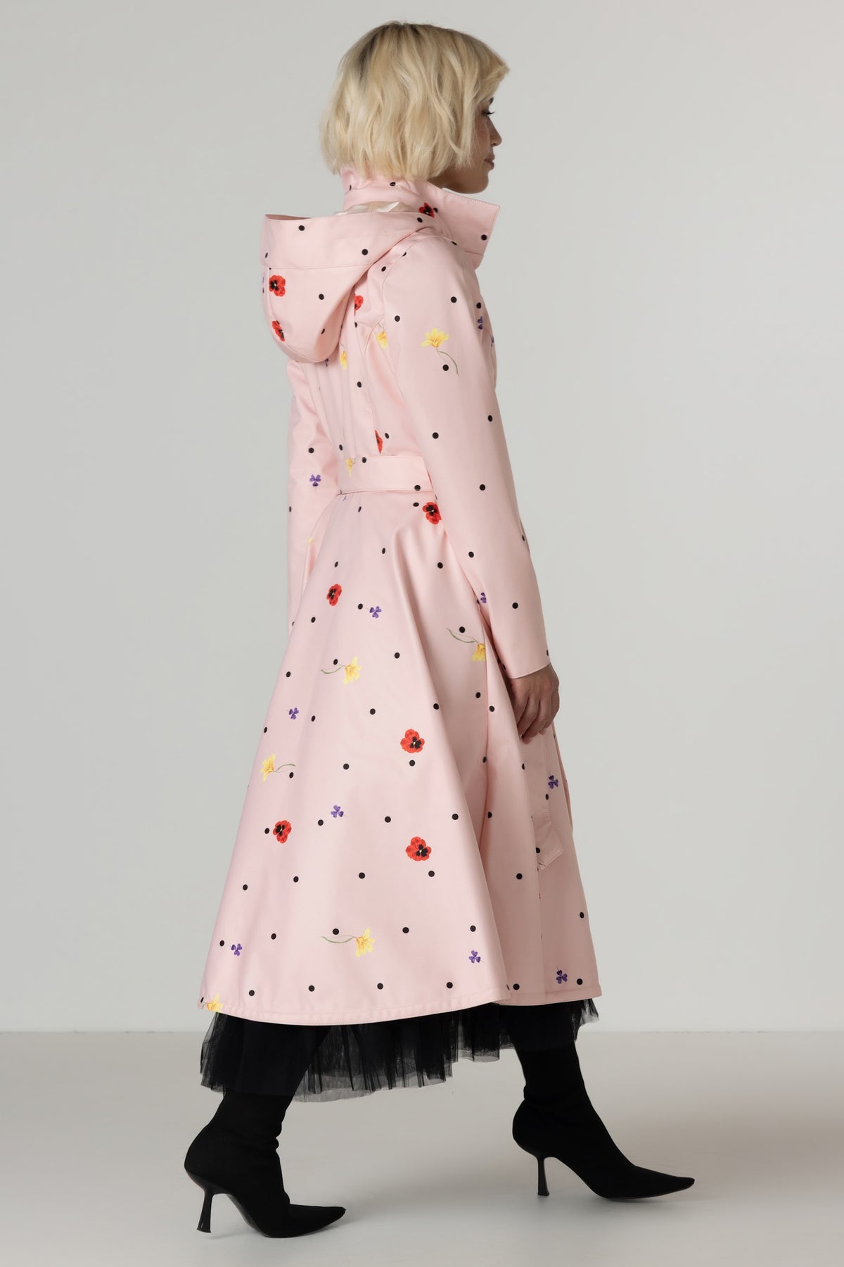 A model shown from the side wearing RainSisters light pink hooded raincoat Cotton Candy with a colorful floral and polka dot print, paired with black tights and black heeled ankle boots, against a neutral light gray studio background.