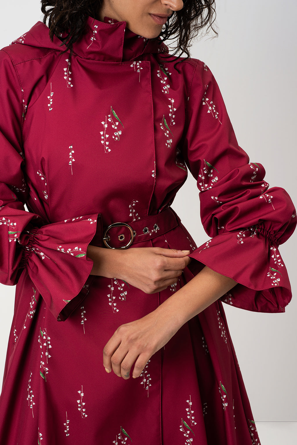 A close-up of a model wearing RainSisters deep red waterproof coat Crimson Lily with white floral print, featuring a high collar, ruffled sleeves, and a matching belt.