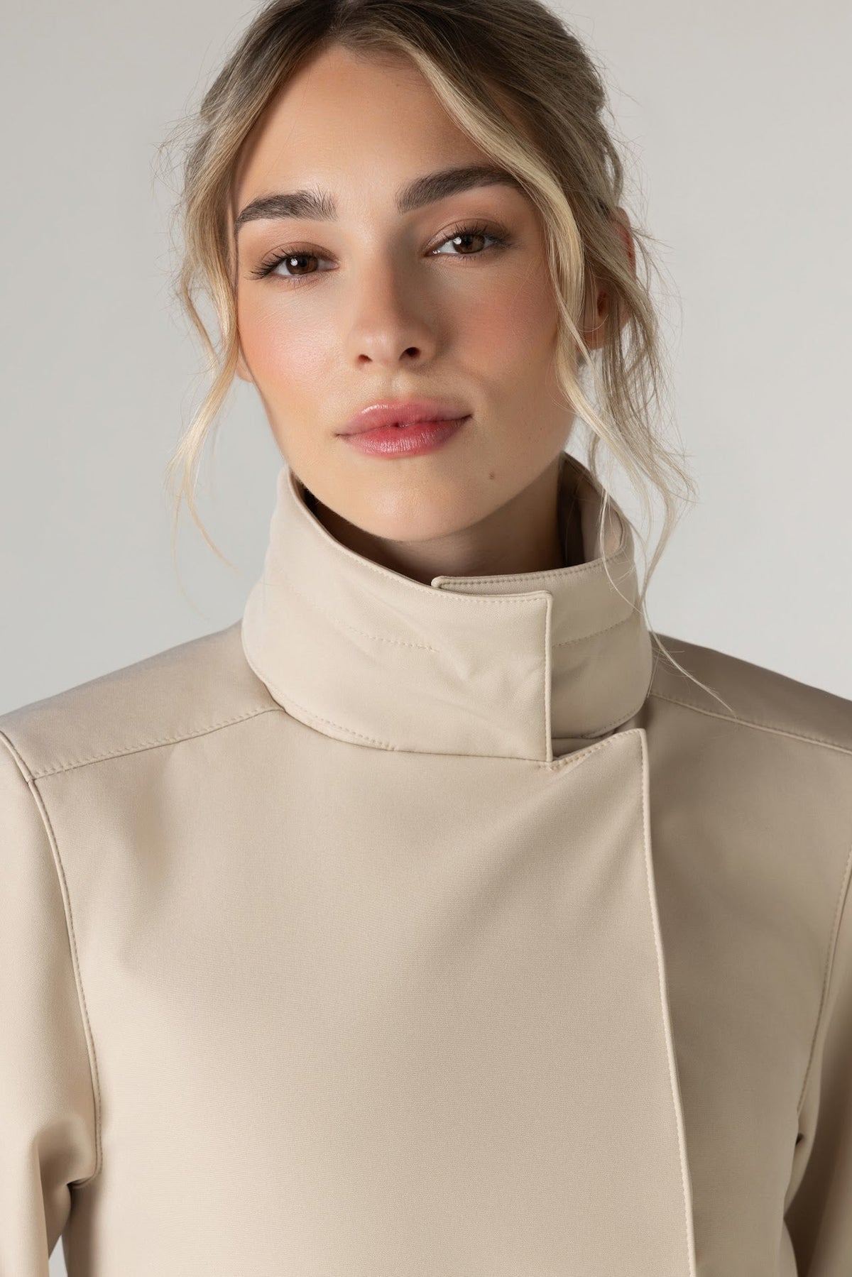 A woman wearing RainSisters beige high collar coat Desirable Bow, shown from the chest up against a plain light gray studio background.