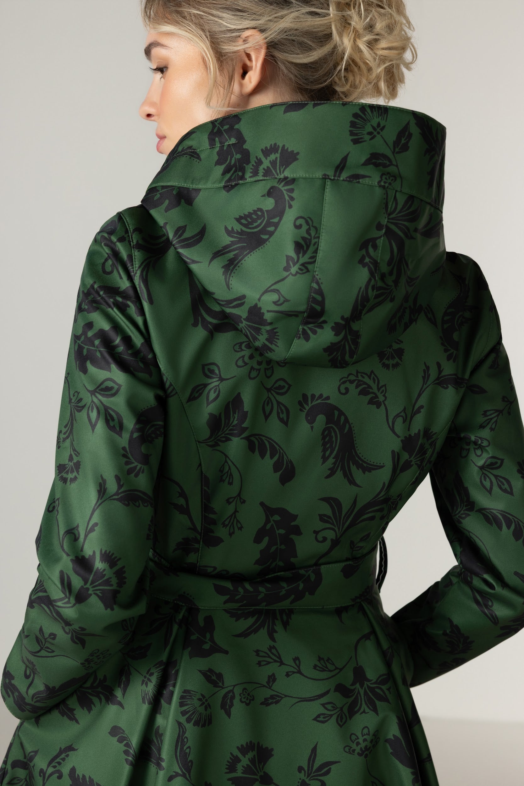A woman shown from the back wearing RainSisters dark green belted coat Forest Flower with black damask-style floral and bird pattern, photographed against a plain light gray studio background.