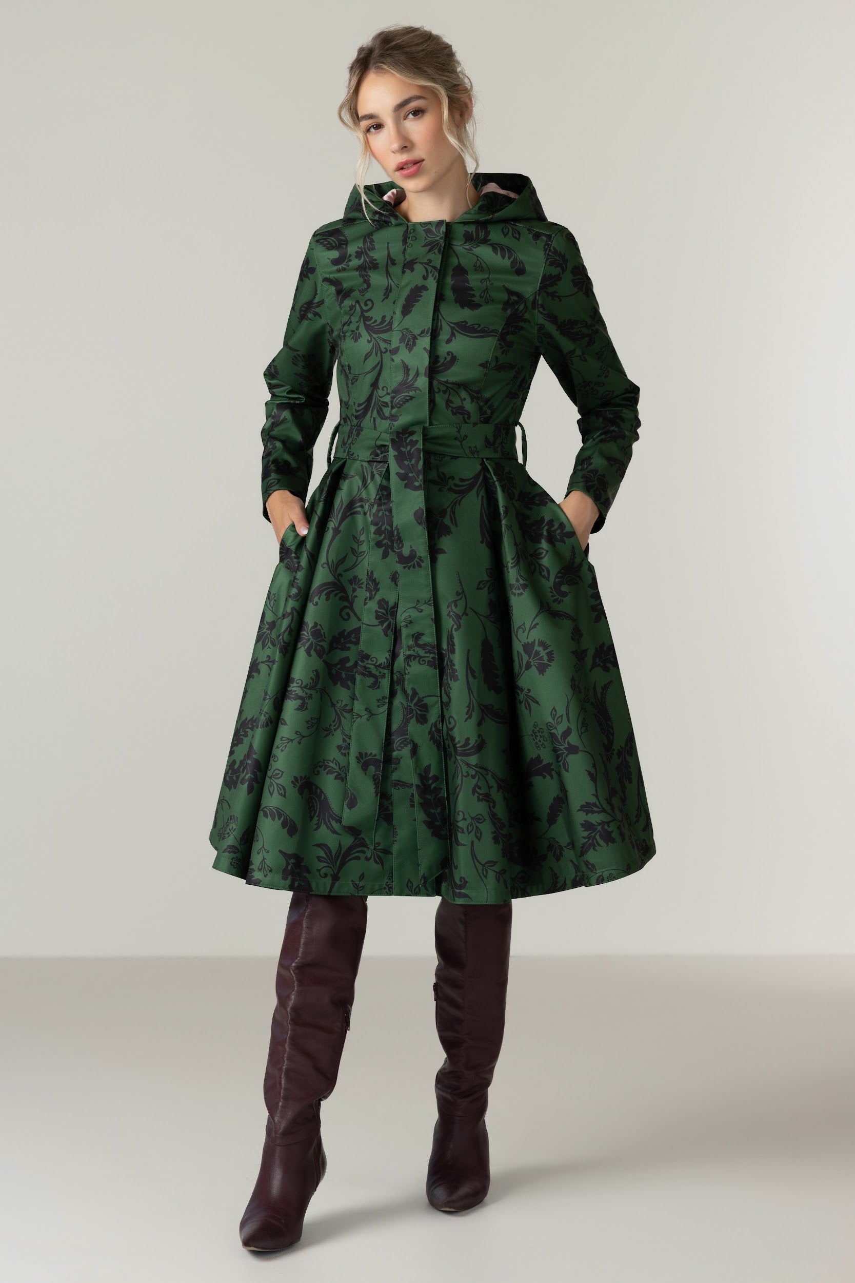 A woman modeling RainSisters dark green hooded coat Forest Flower with a black floral pattern, paired with dark brown over-the-knee boots, against a neutral light gray studio background.