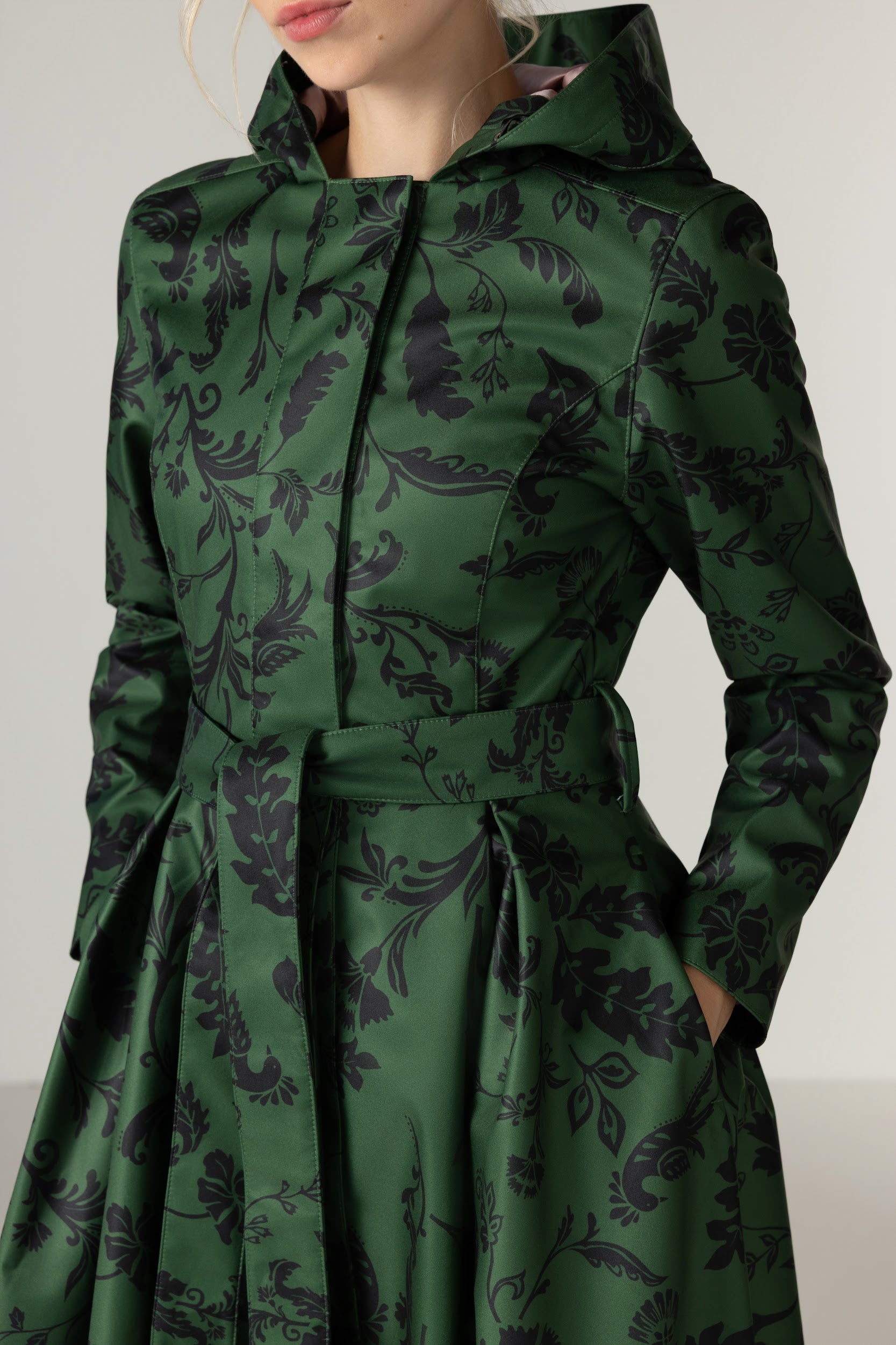 A close-up of a model wearing RainSisters green hooded raincoat Forest Flower with a black floral pattern, featuring a matching waist tie belt and long sleeves.