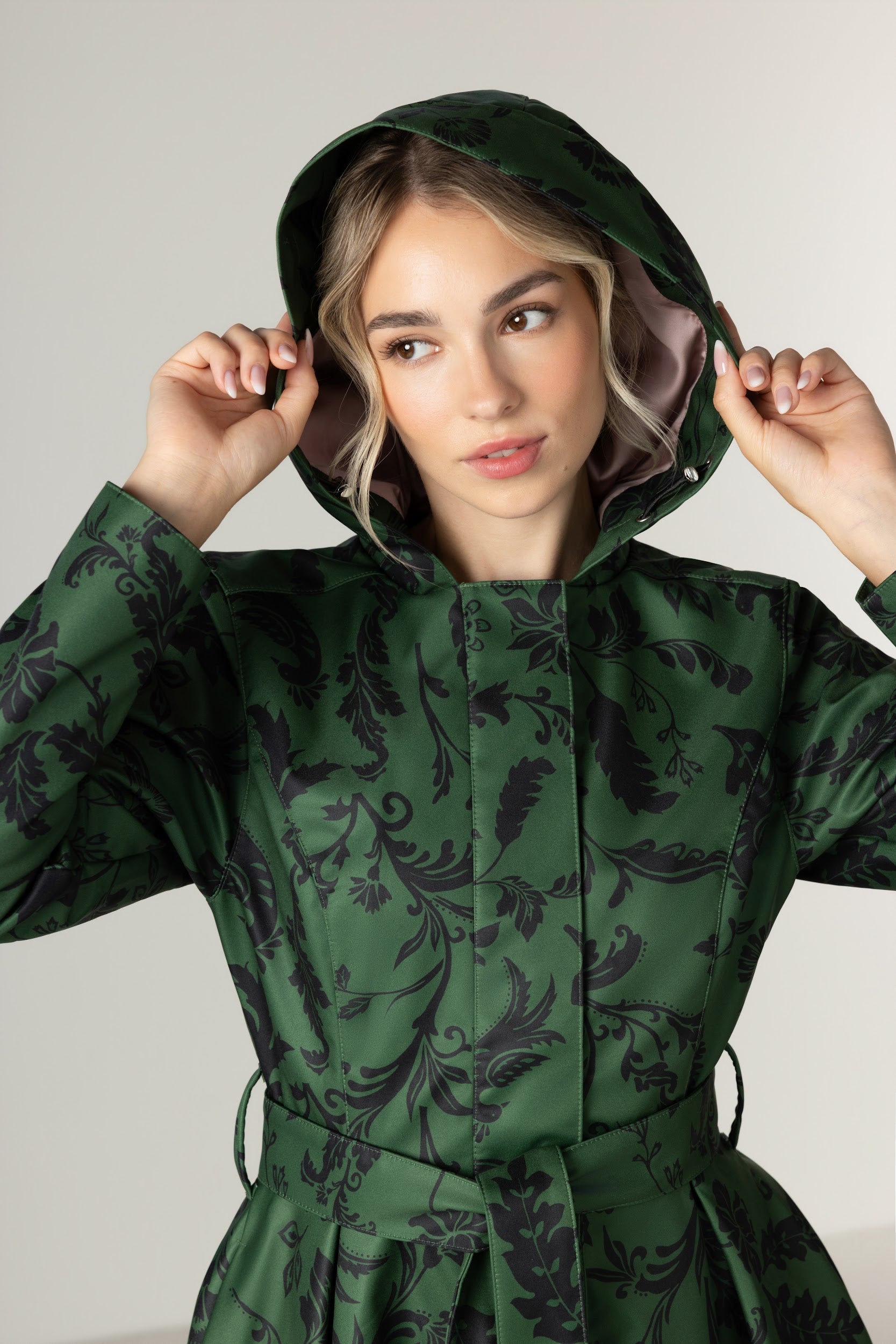 A woman wearing RainSisters dark green hooded raincoat Forest Flower with a black floral pattern, holding the hood up around her face against a plain light gray background.
