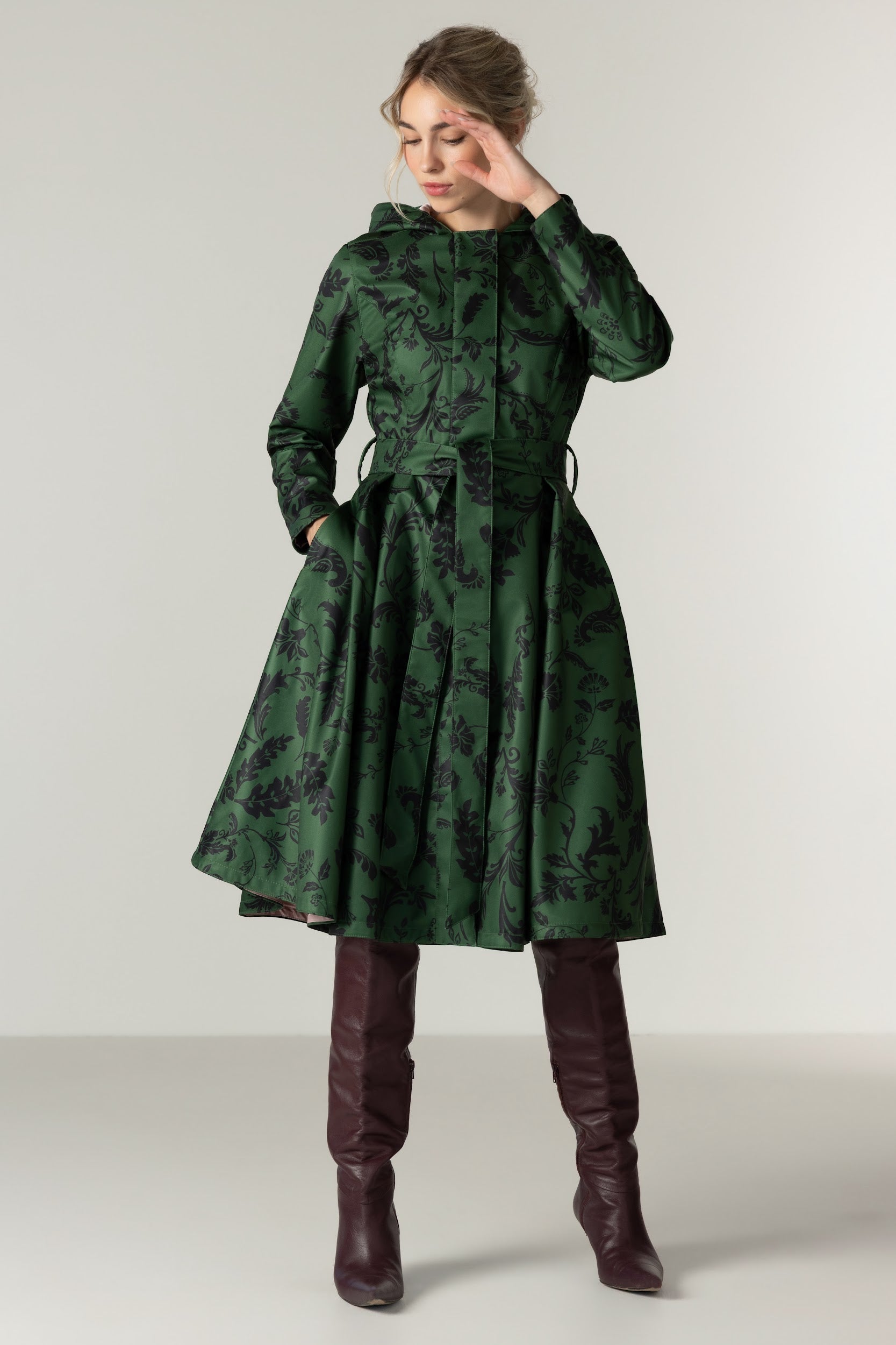 A woman models RainSisters dark green hooded waterproof coat Forest Flower with a black floral pattern, paired with dark brown knee-high boots, against a neutral light gray studio background.