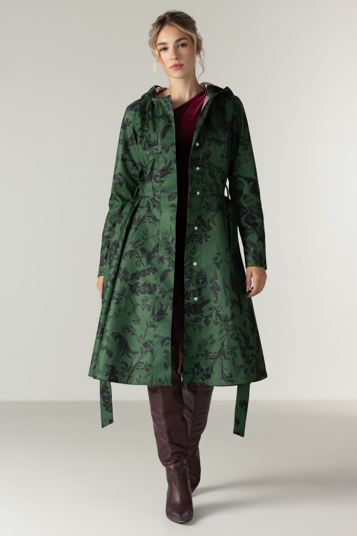 A woman models RainSisters long green hooded raincoat Forest Flower with a dark floral damask pattern, worn over a burgundy top and dark brown over-the-knee boots, against a neutral studio background.
