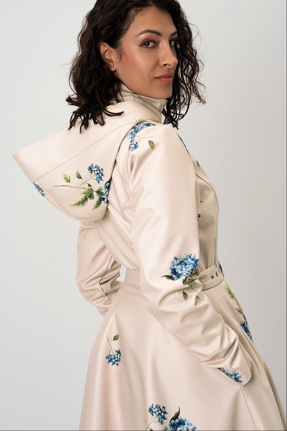 A woman wearing RainSisters cream-colored hooded trench coat Gentle Bloom with blue and green floral hydrangea print, photographed against a light gray studio background.