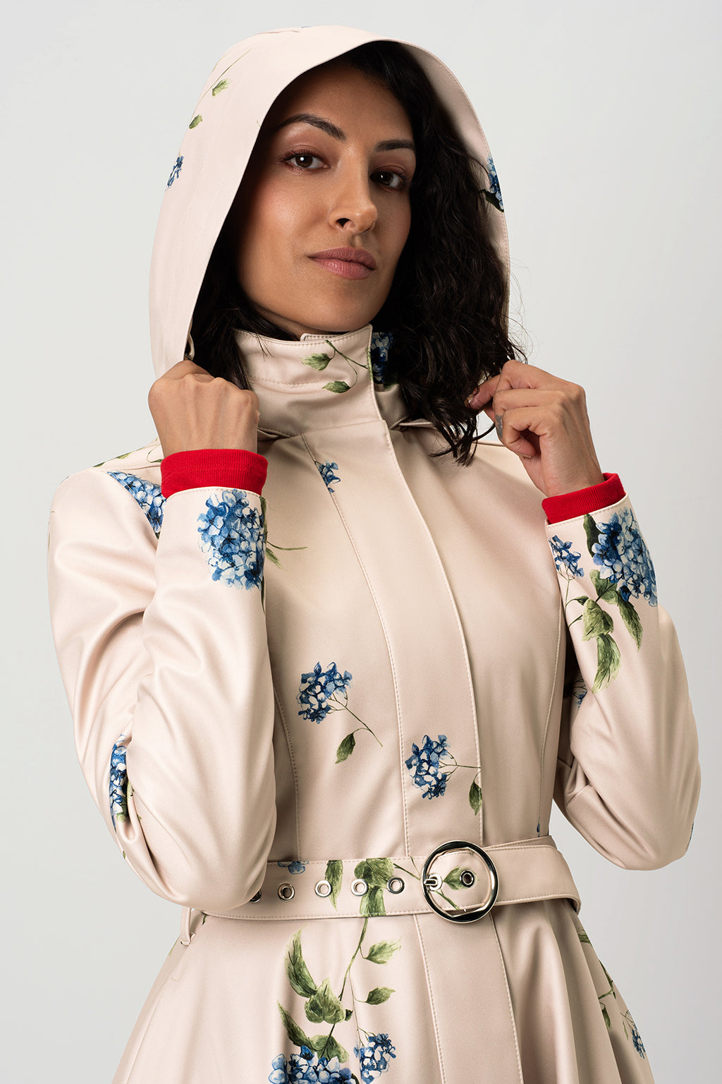 A close-up of RainSisters beige trench coat Gentle Bloom with blue floral prints and a matching belt with a silver buckle and hood on, shown on a model against a light gray background.