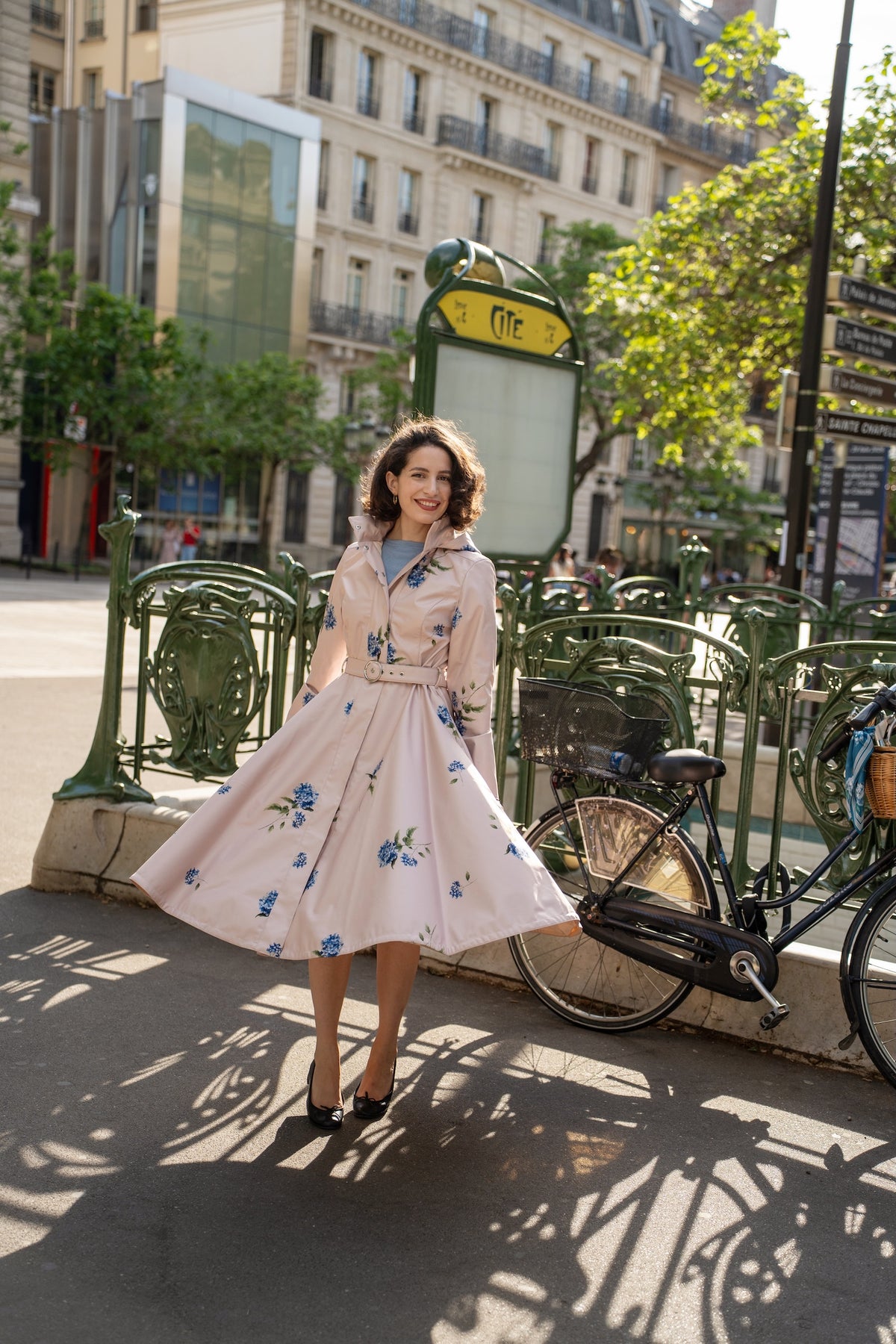 A woman stands outdoors wearing RainSisters long, light blush pink raincoat Gentle Bloom with blue floral print