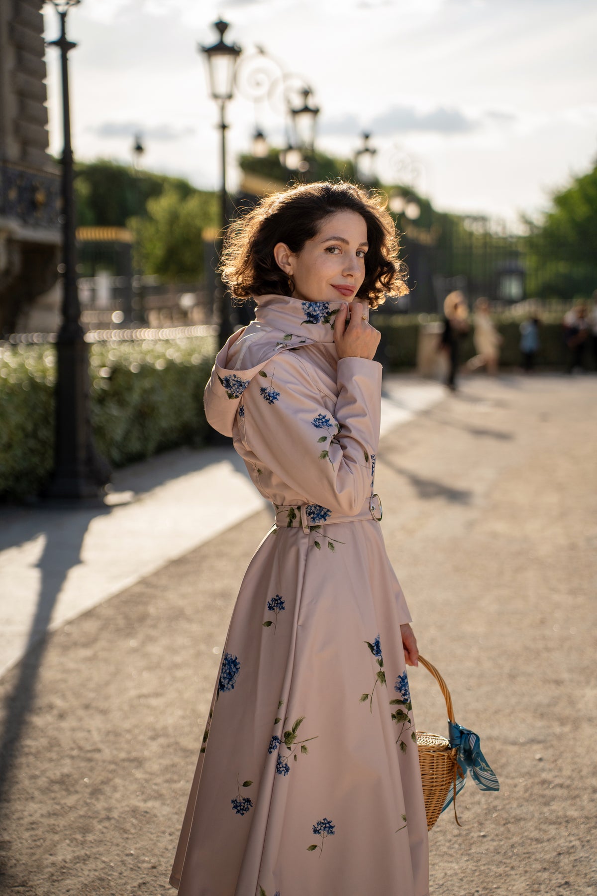 A woman stands outdoors in a sunlit park-like setting wearing RainSisters long, light blush pink raincoat Gentle Bloom with blue floral print and holding a woven basket.