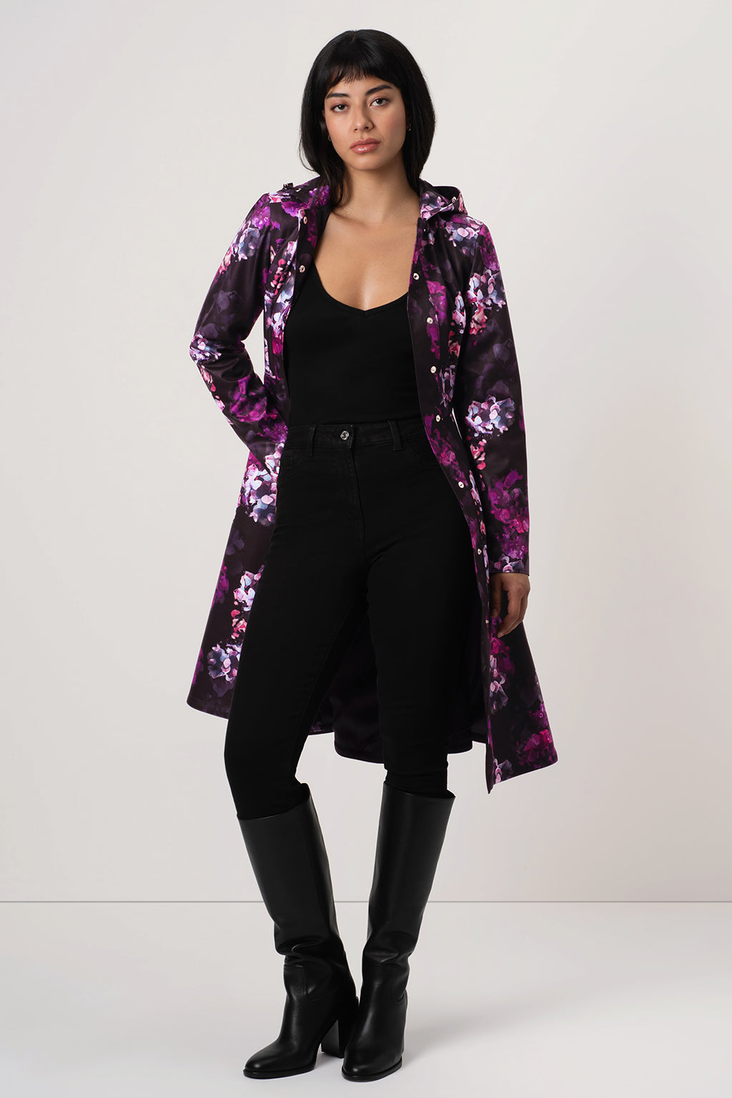 A model stands against a light gray studio background wearing RainSisters long black hooded raincoat Hortense with a vibrant purple and pink floral pattern, styled over a black outfit and tall black boots.