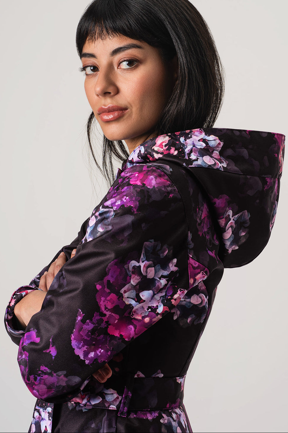 A woman wearing RainSisters black hooded jacket Hortense with a vibrant purple and pink floral print, photographed against a neutral light gray background.