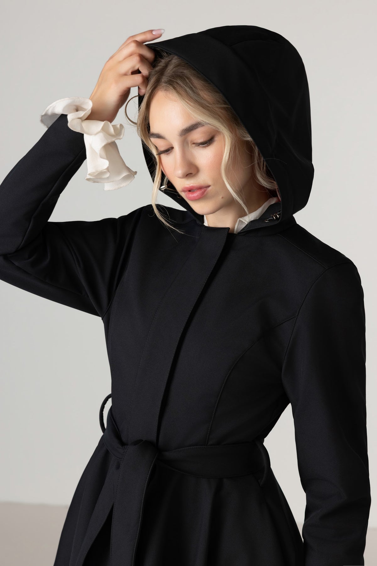 A woman wearing RainSisters black hooded waterproof coat Midnight Black with a matching waist tie, styled with a white ruffled collar detail, set against a plain light gray background.