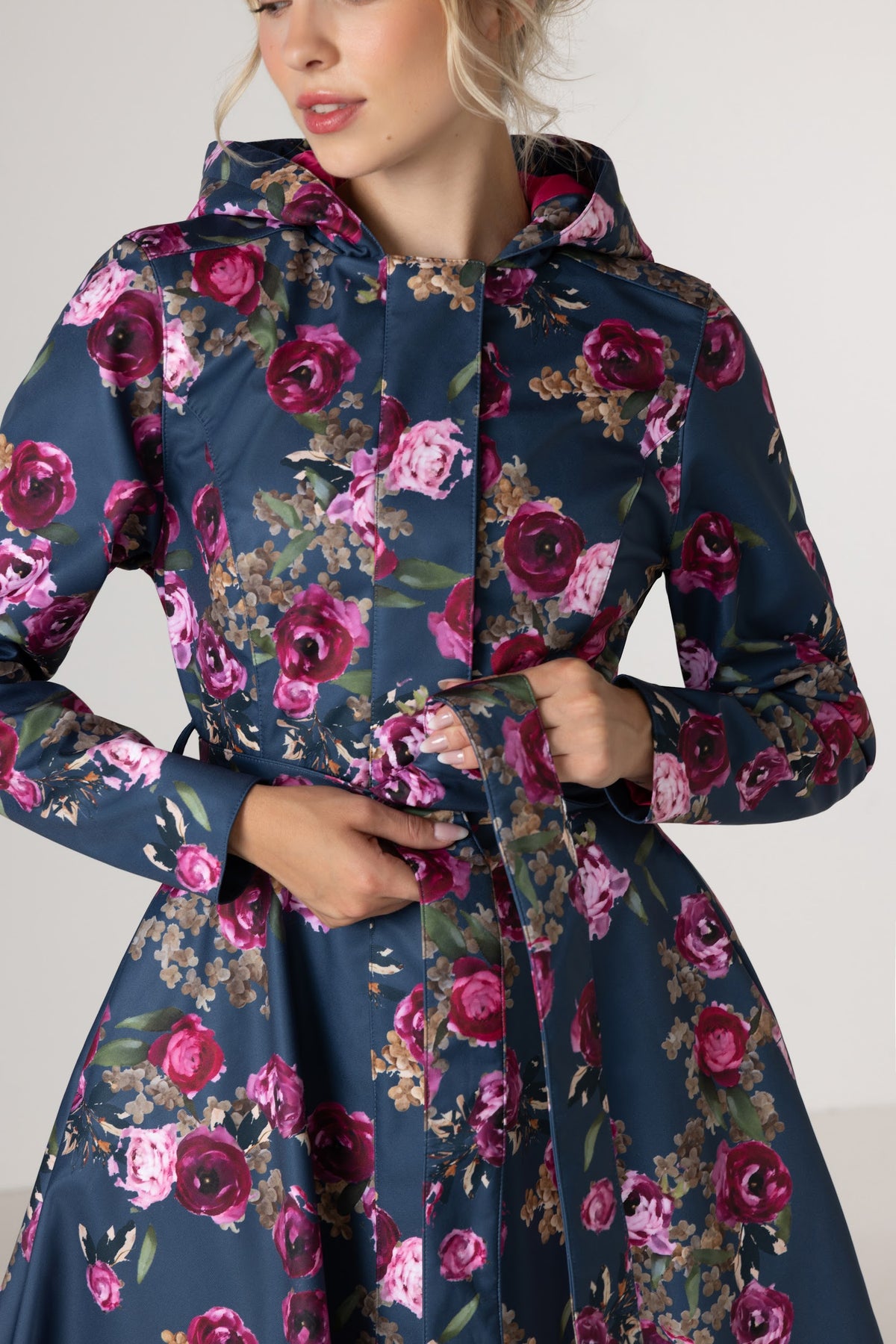 A woman wearing RainSisters navy blue hooded raincoat Misty Rose with a pink and purple floral print, featuring a matching waist tie belt, shown against a plain light gray background.