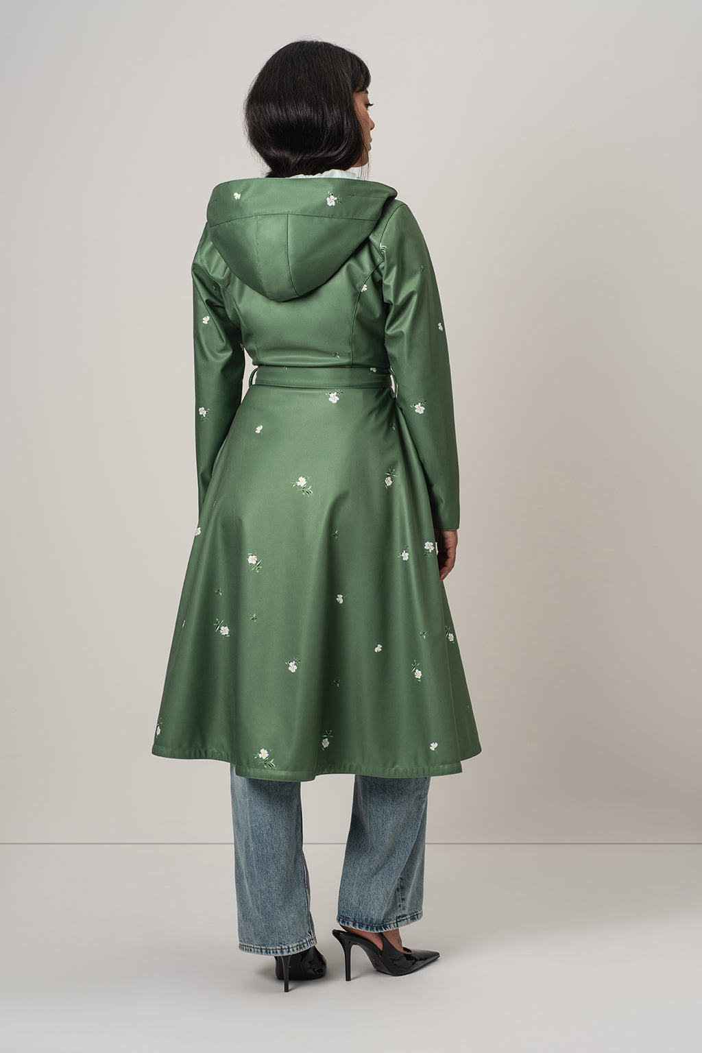 A woman viewed from the back wearing RainSisters green hooded raincoat Primrose in Bloom with small white floral prints, paired with blue jeans and black heels, against a plain light gray studio background.