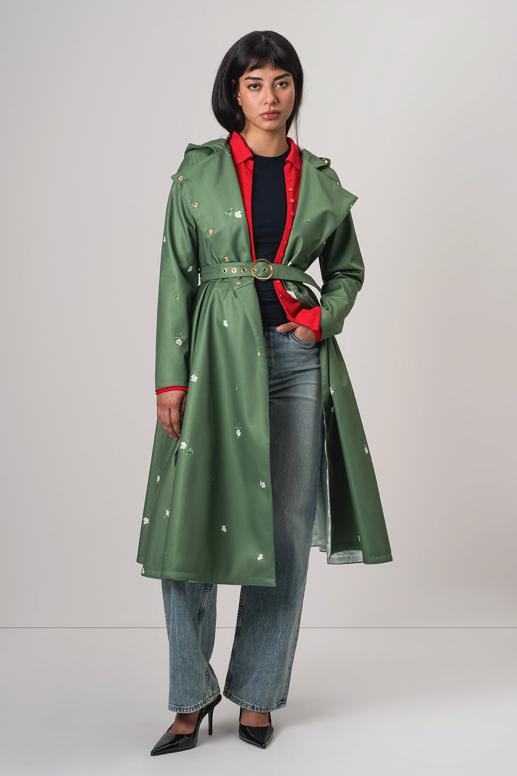 A model wearing RainSisters green belted trench coat Primrose in Bloom with small white floral prints, layered over a red collared shirt, black top, blue jeans, and black heels, photographed against a light gray studio background.