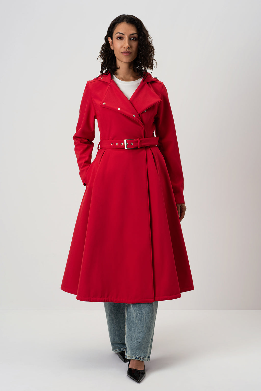 A woman modeling RainSisters bright red mid-length trench coat Queen of Hearts with a matching waist belt, worn over a white top and light blue jeans, against a plain light gray studio background.