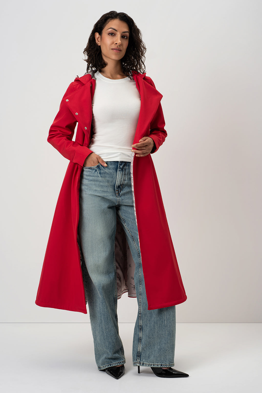 A woman modeling RainSisters long red trench coat Queen of Hearts over a white top and light blue jeans, standing against a plain light gray studio background.