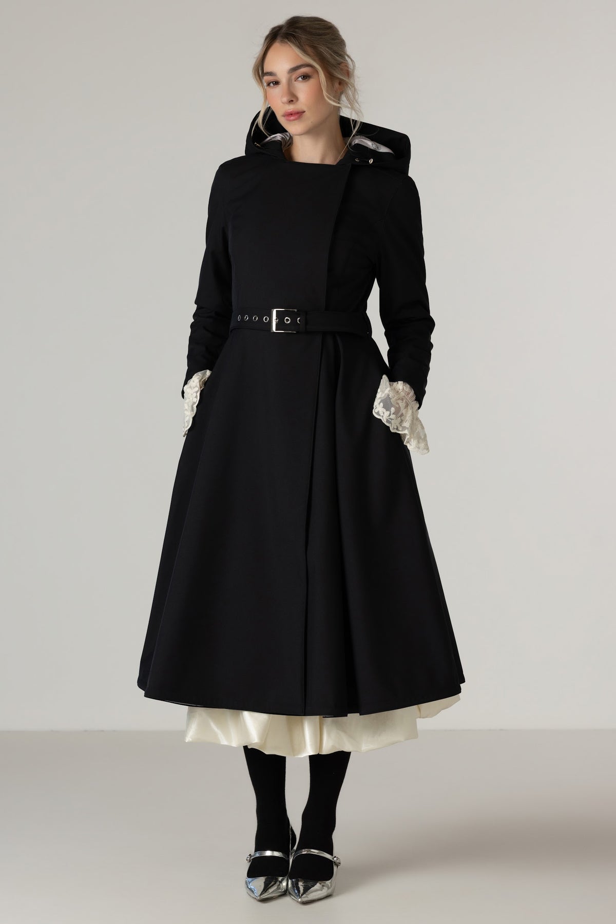 A woman models RainSisters black longline coat Queen of Spades with a matching waist belt, styled white lace cuffs, over a cream dress, black tights, and silver shoes against a plain light gray studio background.