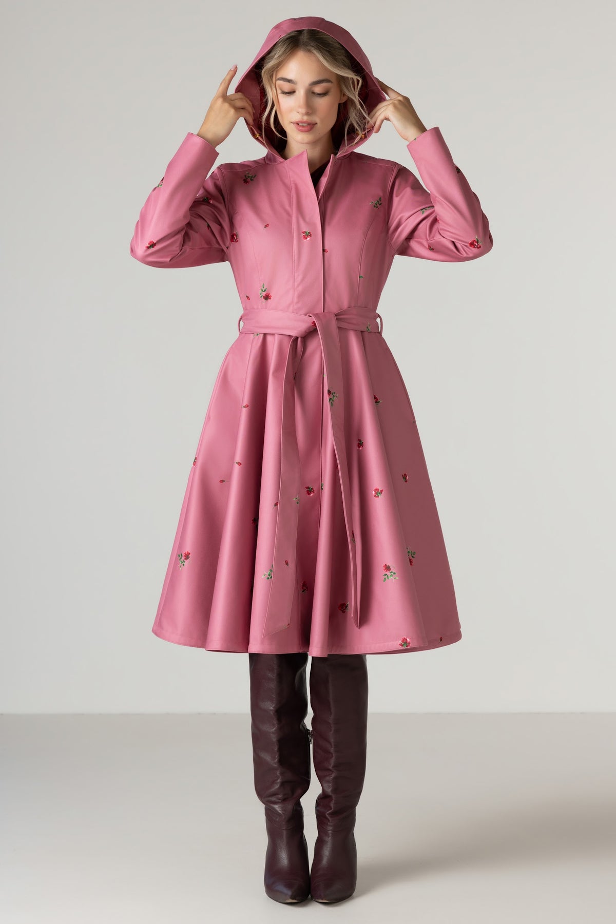 A woman wearing RainSisters pink hooded raincoat Raspberry Sorbet with a matching waist tie belt and small rose flower print, paired with dark brown knee-high boots, standing against a light gray studio background.