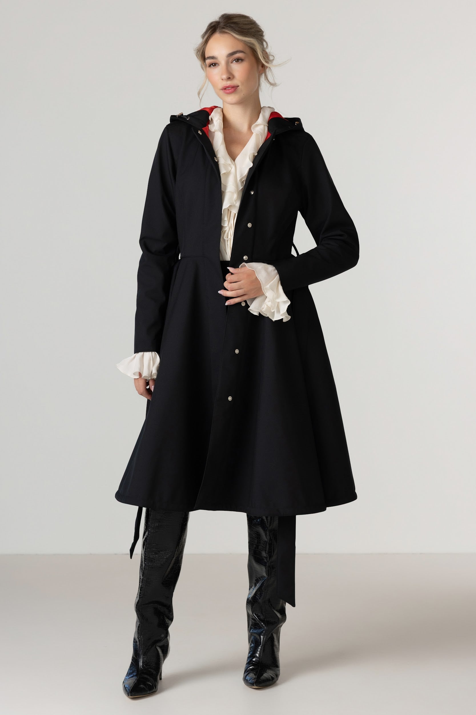 A model wearing RainSisters long black coat Raven Red with a red inner lining, layered over a white ruffled blouse, black pants, and black boots, standing against a light gray studio background.