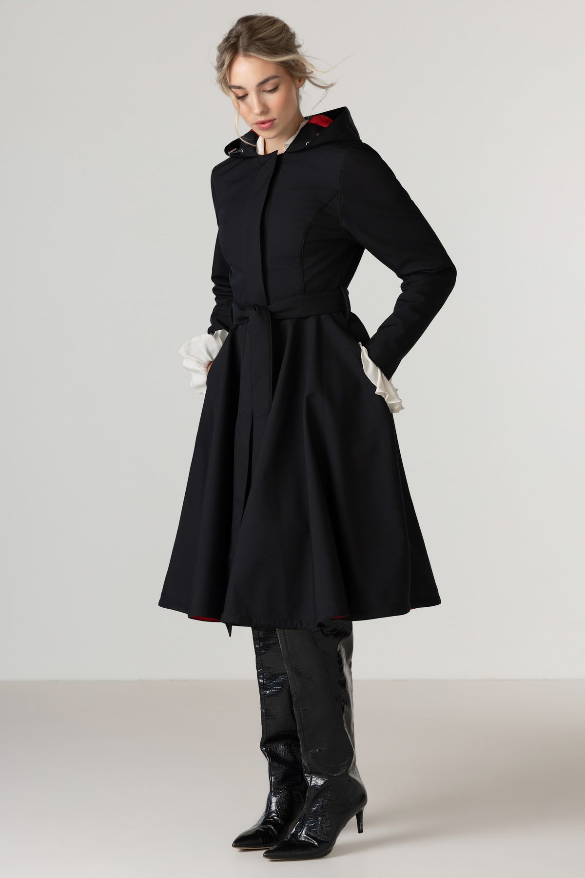 A woman modeling RainSisters black hooded raincoat Raven Red with a belted waist and black knee-high boots against a plain light gray studio background.
