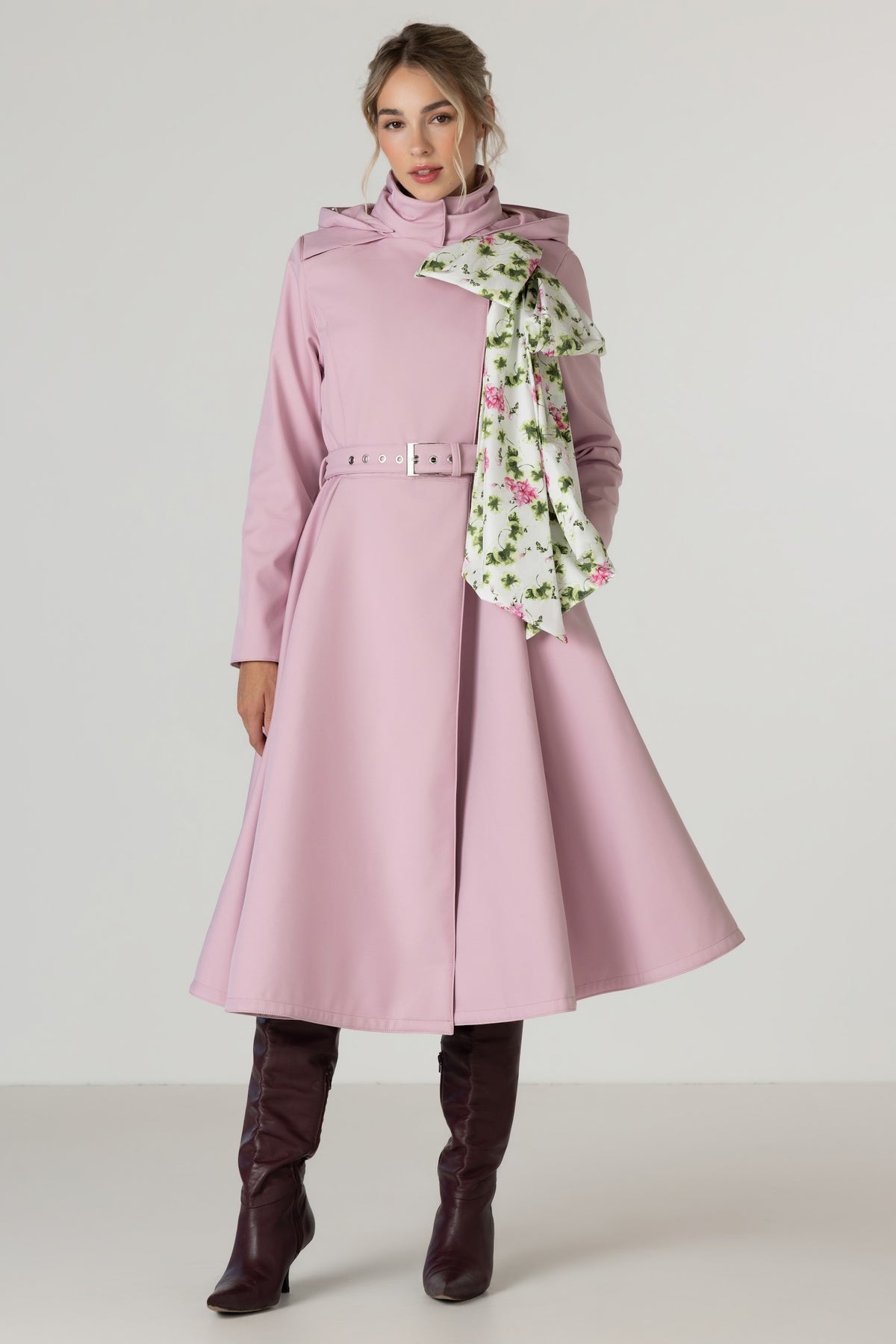 A woman models a light pink mid-length coat Rosie Bow by RainSisters. The coat has high collar, belt, and flared skirt, green and white detachable floral scarf. Styled with dark brown knee-high boots against a plain light gray studio background.