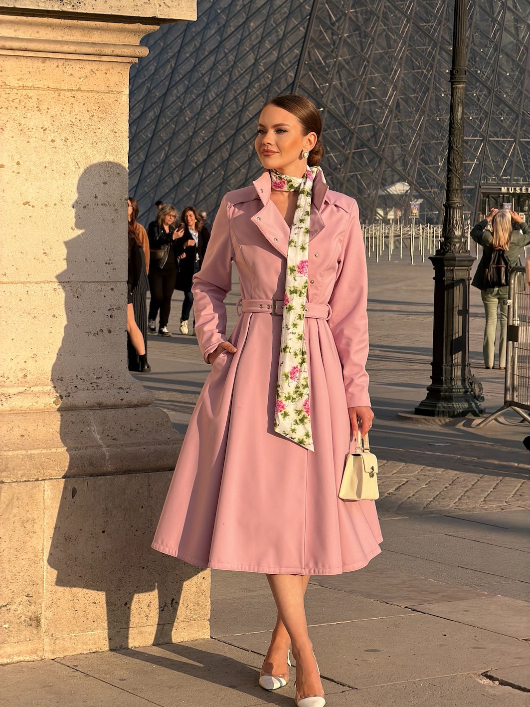 A woman stands outdoors in front of the Louvre Pyramid in Paris, wearing RainSisters light pink knee-length trench coat Rosie Bow with a matching belt, a floral print neck scarf, and holding a small cream handbag.