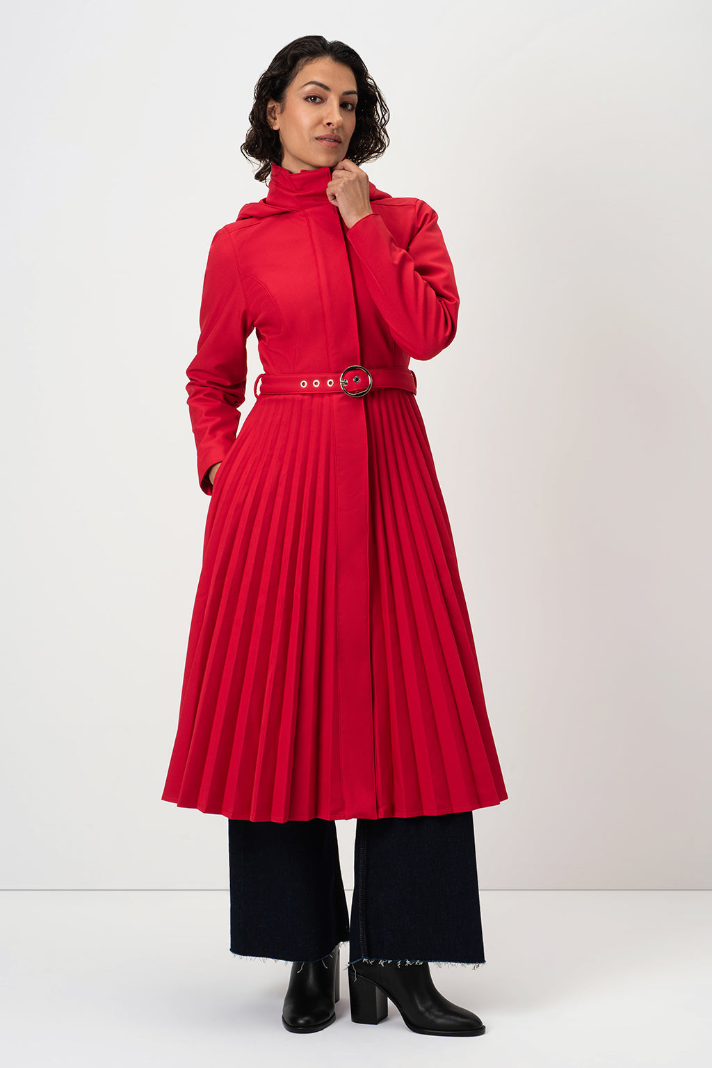 A model wearing RainSisters bright red long-sleeve coat Scarlet Waltz with a pleated skirt and a matching belt, paired with black wide-leg pants and black boots, against a plain light gray studio background.