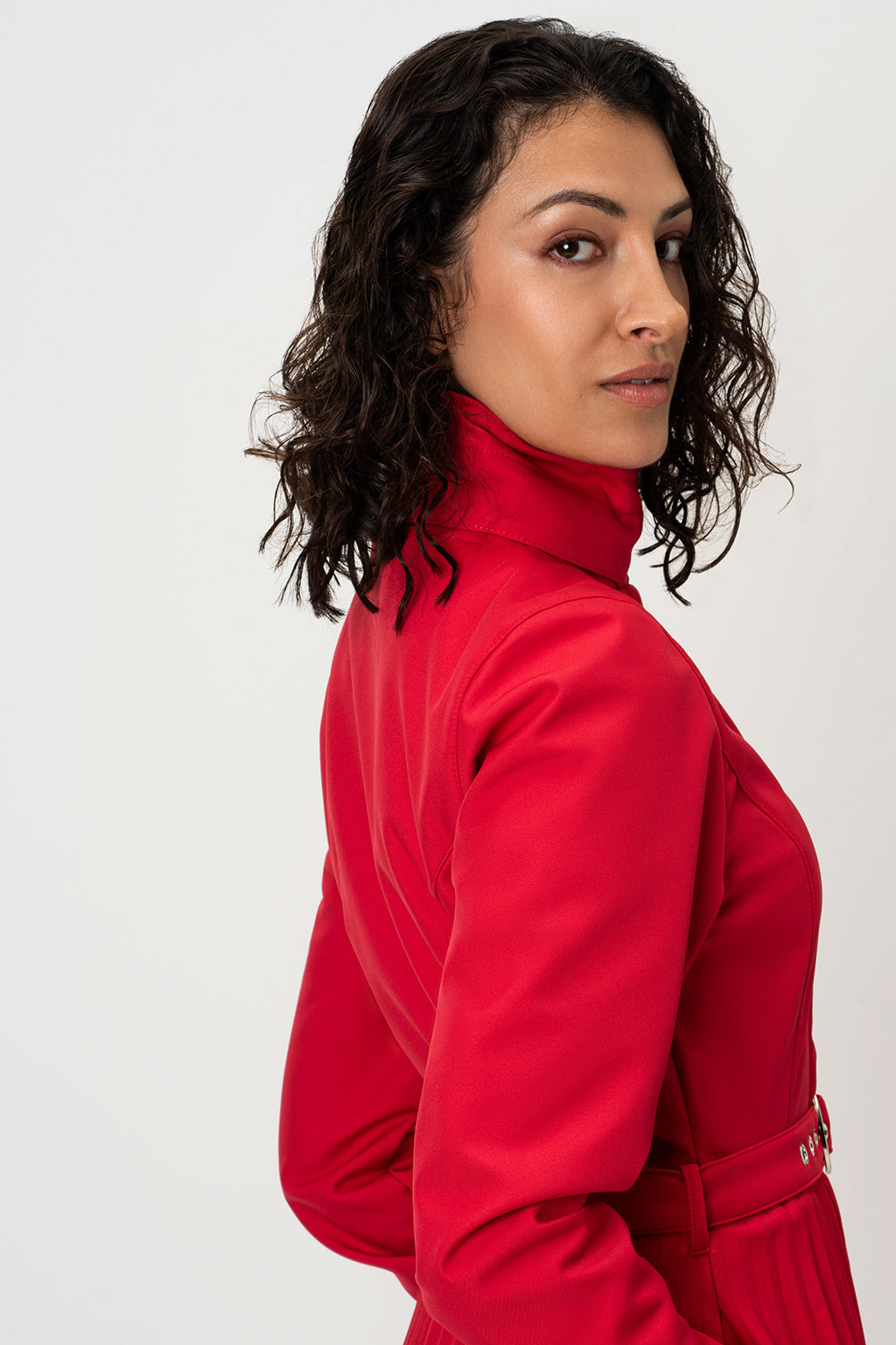 A woman with dark curly hair is shown from a side-back angle wearing RainSisters bright red high-neck raincoat Scarlet Waltz against a plain light gray studio background.