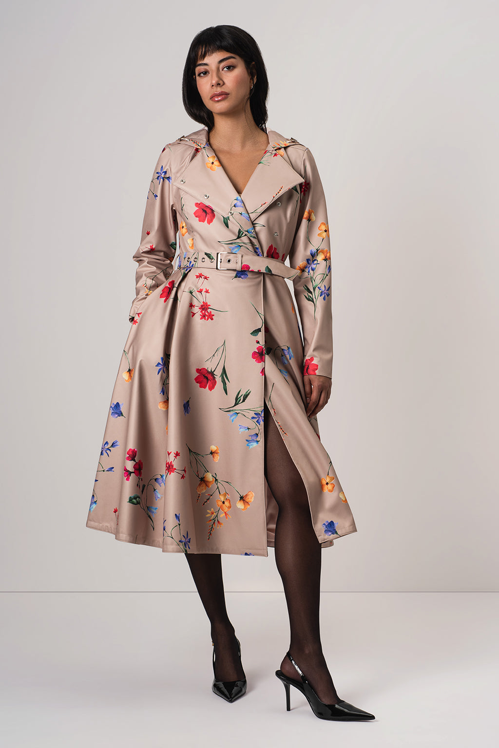 A woman models RainSisters beige satin-like trench coat Spring Bloom with a colorful floral print, featuring a matching belt and a thigh-high slit, paired with black tights and heels against a light gray studio background.
