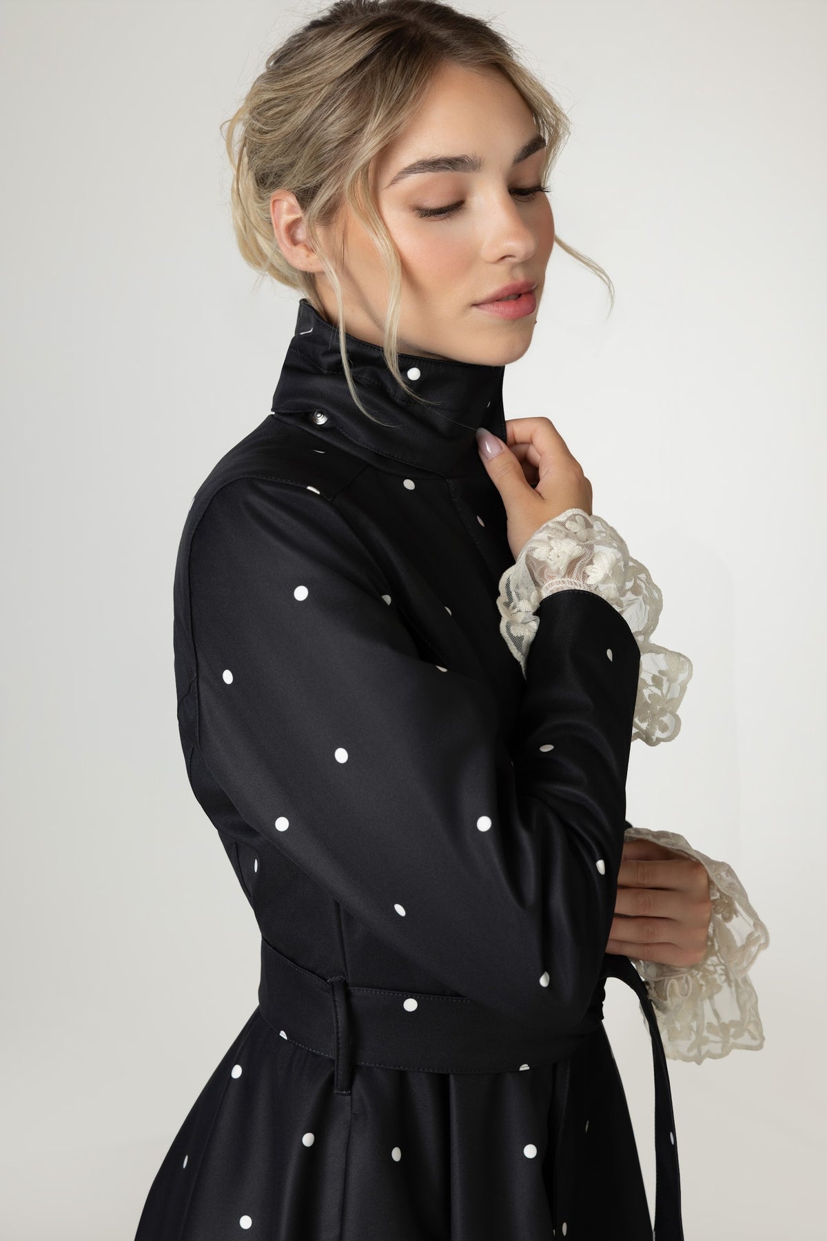 A woman wearing RainSisters black long-sleeve raincoat Timeless Polka Dots with white polka dots, shown in a side profile against a light gray studio background.