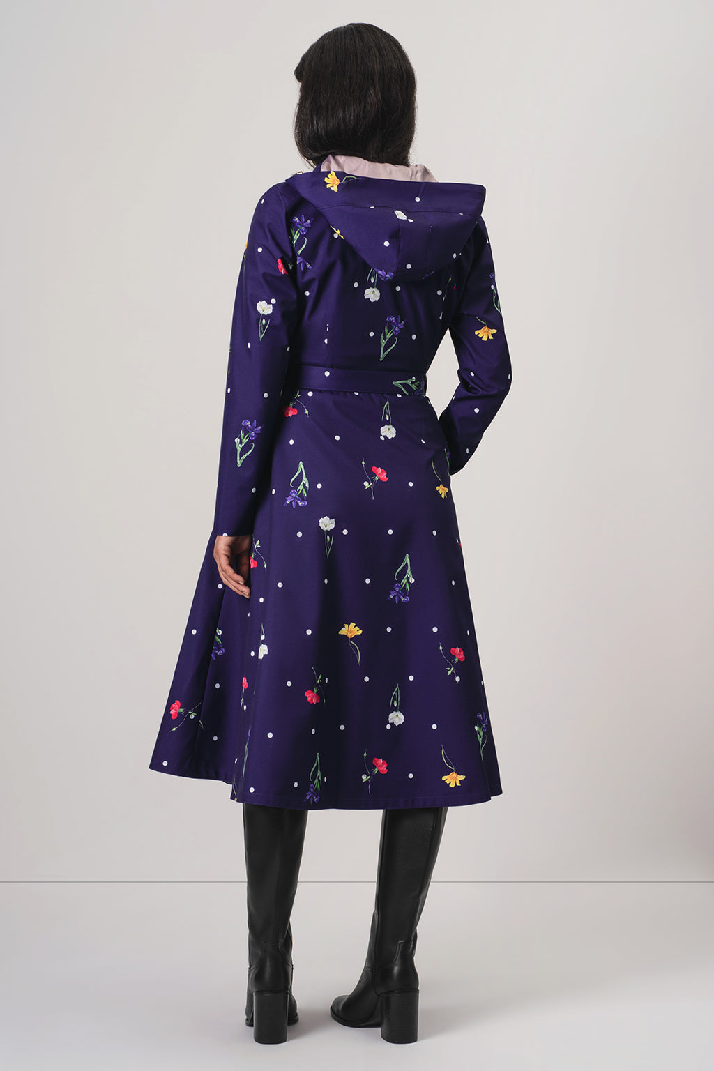 A model viewed from the back wearing RainSisters dark purple hooded coat Violet Bloom with a colorful floral and polka dot pattern, paired with black knee-high boots against a light gray studio background.