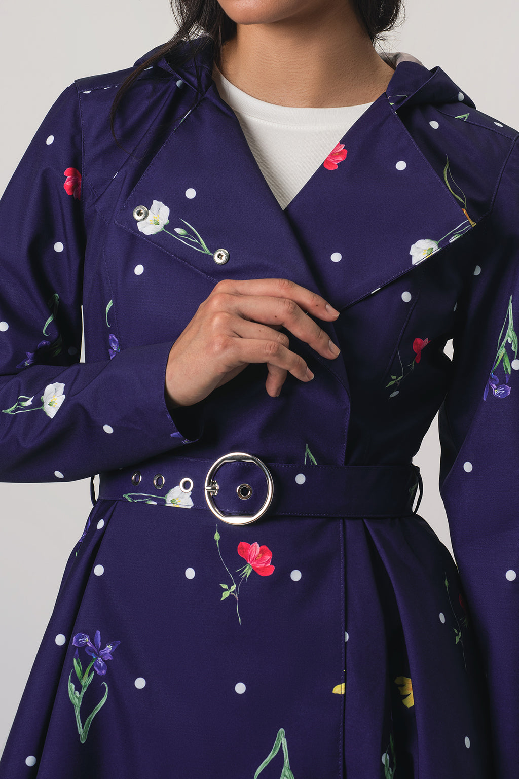 A close-up of a model wearing RainSisters purple hooded raincoat Violet Bloom with a white polka dot pattern and colorful floral print, featuring a matching belt with a silver buckle.