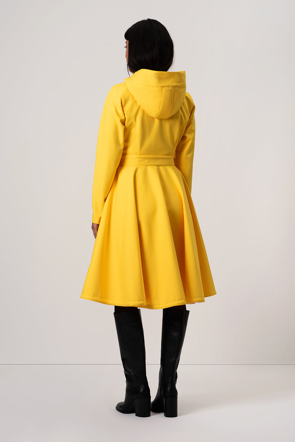 A model viewed from the back wearing RainSisters bright yellow hooded A-line coat Yellow Sun with a tie waist, paired with black knee-high boots, against a plain light gray studio background.