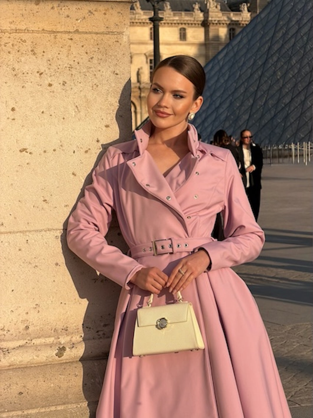 A woman wearing RainSisters dusty pink double-breasted trench coat Rosie Bow with a matching belt and holding a cream-colored handbag, posing outdoors in front of a stone wall and modern glass structure.