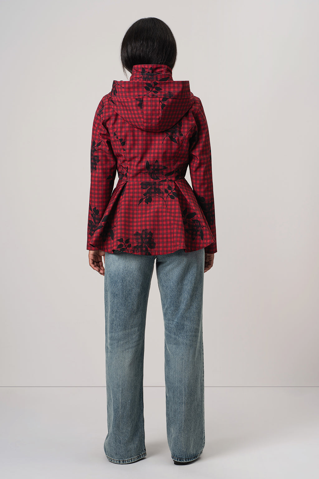 A model viewed from the back wearing RainSisters red and black checkered hooded jacket Crimson Heart with a black floral pattern, paired with light blue jeans, against a plain light gray background.